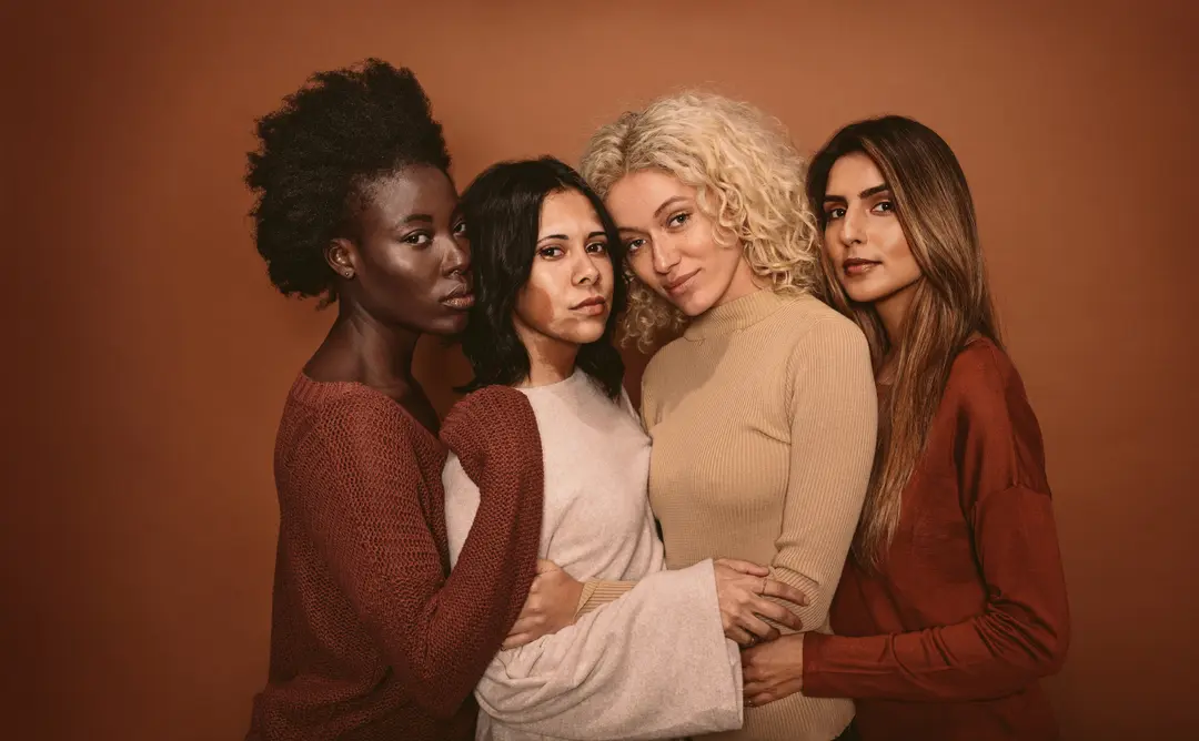Quatro mulheres abraçadas, vestindo tons de terra, olhando para a câmera.